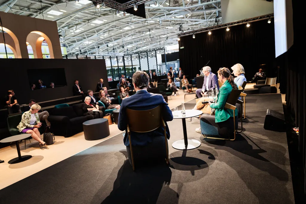 Ein weitwinklig von hinten fotografierter Mann sitzt mit mehreren Rednern und Rednerinnen auf einem Podium einer Panel-Diskussion einer Messe. Foto von William Veder · Eventfotografie | Eventfotograf aus Berlin