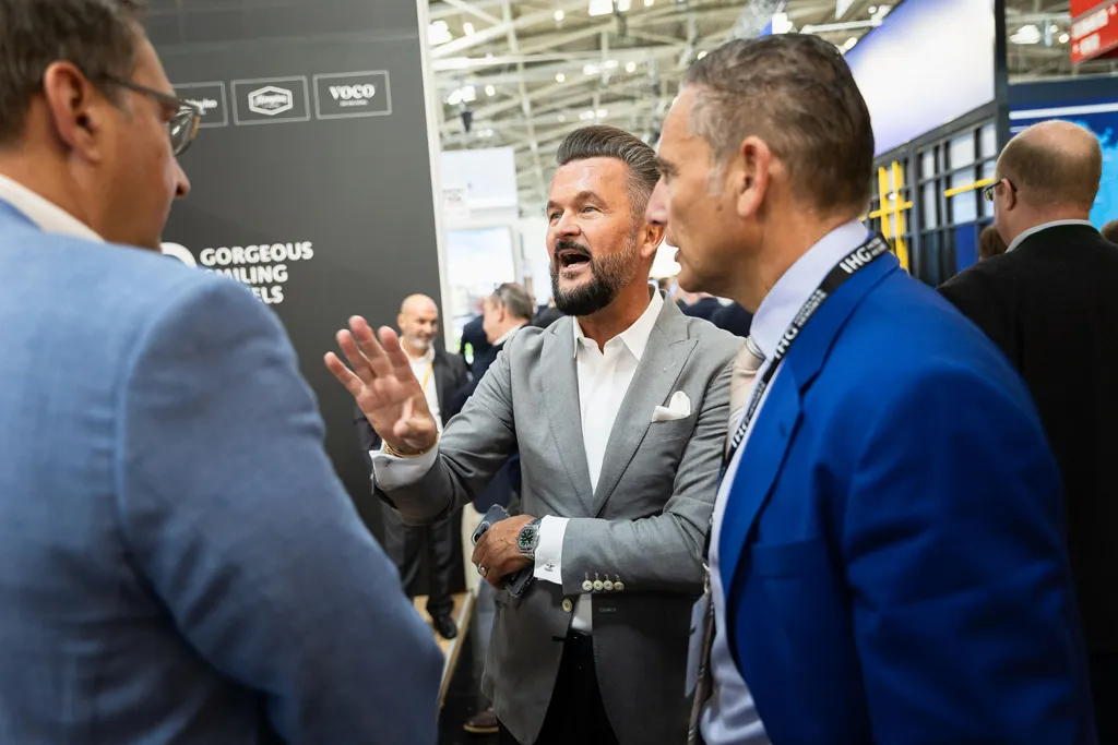Drei Hotelmanager in Anzügen auf der Immobilien-Messe Expo-Real in München stehen zu einem Gespräch im Kreis, während die mittige Person zu einem Redebeitrag die rechte Hand hebt.