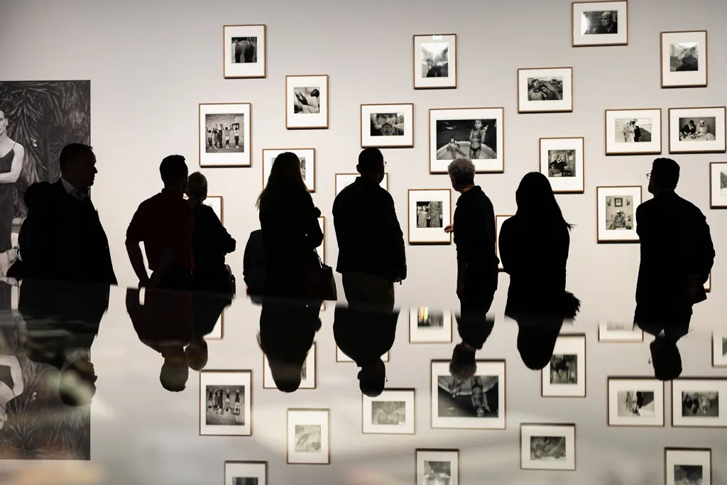 Eine Menschengruppe in einer Fotoausstellung, die sich in einer Glasvitrine spiegelt, sodass die Oberkörper auch die Unterkörper bilden.