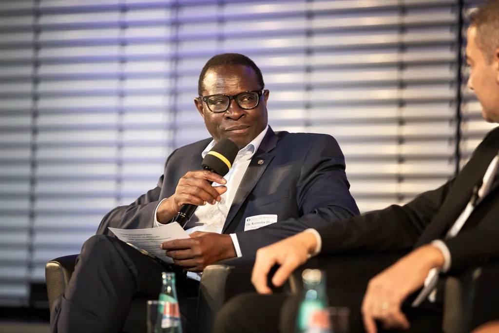 Großaufnahme von Dr. Karamba Diaby, der zum diesem Zeitpunkt Mitglied des Deutschen Bundestages ist und auf einer Konferenz auf einem Podium in einem Sessel sitzt und einen Redebeitrag in ein Mikrofon spricht.