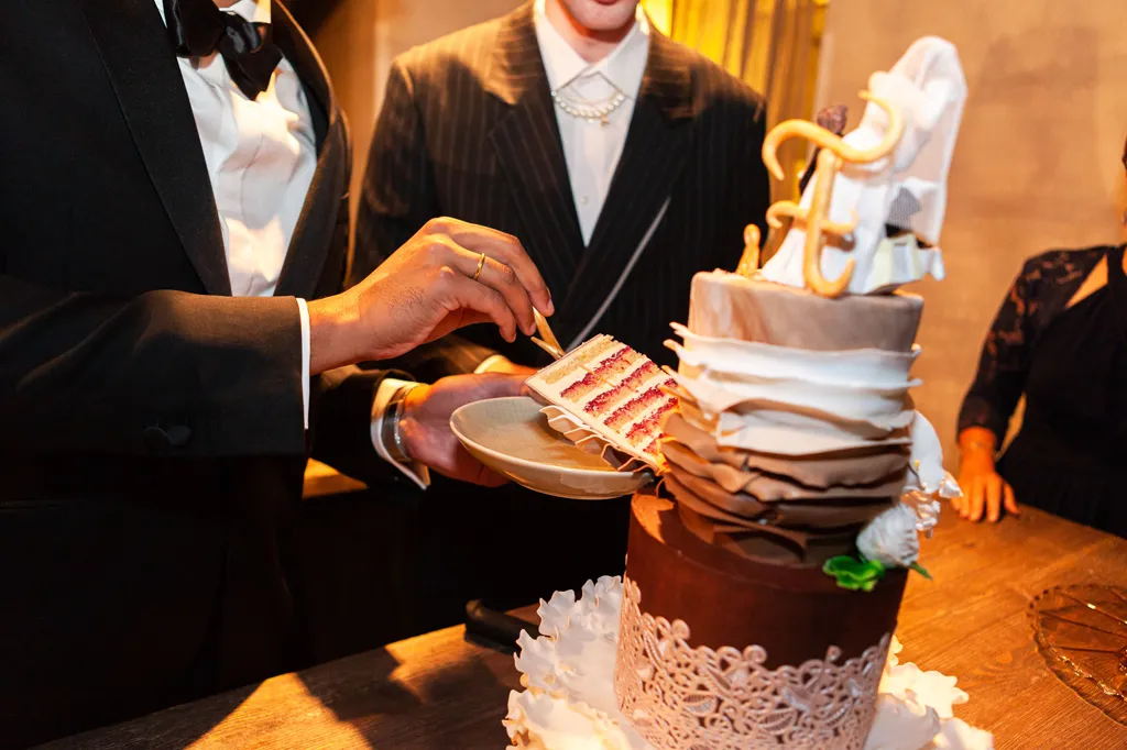 Zwei Männer ab unterhalb des Kinnes fotografiert stehen vor einer Hochzeitstorte, während die Hand des eines Mannes mit einer Kuchengabel in ein Stück Torte sticht, die sich auf seinem Teller befindet