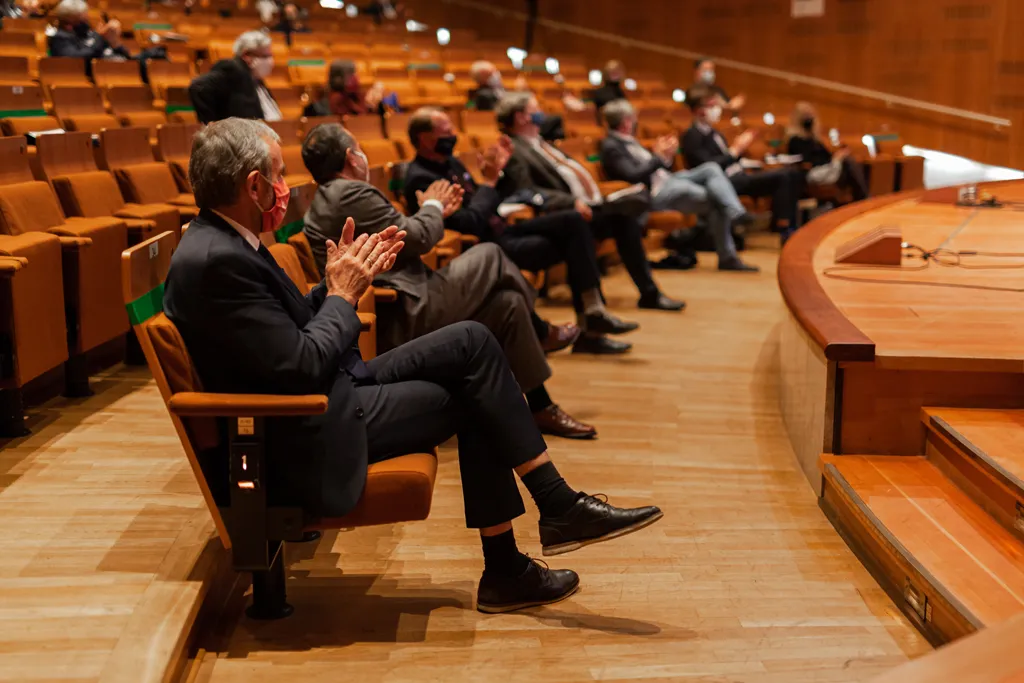 Mehrere Menschen mit Anzügen sitzen in der ersten Reihe einer Veranstaltung in einem holzverkleideten modern anmutenden Auditorium.