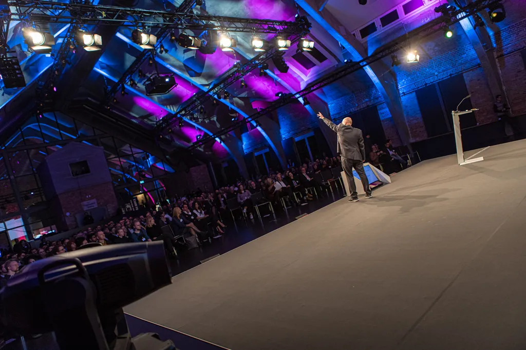 Schräge Weitwinkel-Aufnahme eines Redners von hinten im grauen Anzug, der seinen Arm hebt. Er steht auf einer großen, leeren Bühne und die Beleuchtung des Raumes ist in pink und blau gehalten.