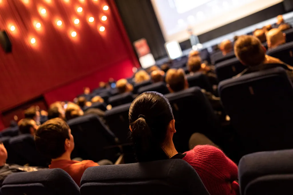 Schräge Weitwinkel-Aufnahme von Personen von hinten, die in einem Kino mit roten Samtwänden und gelber Leuchten-Dekoration sitzen und dem Bühnenprogramm vor einer Leinwand zuschauen, das in der Unschärfe verschwimmt.