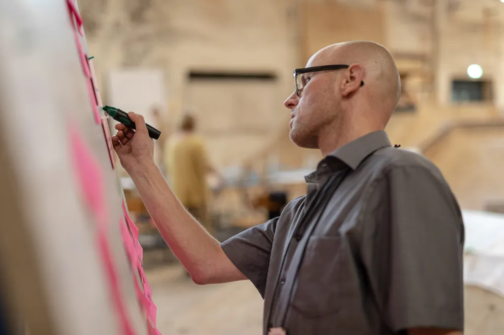 Eine halbnahe Profil-Aufnahme in warmen Tönen zeigt einen Mann mit Glatze und Brille, der mit einem dicken schwarzen Marker an ein Flipchart schreibt.