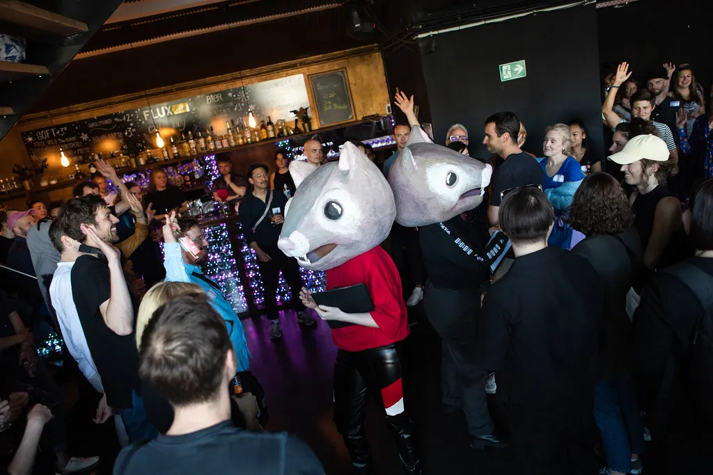 Zwei Personen mit Hamster-Masken aus Pappmaché auf dem Kopf und Klemmbrettern in der Hand stehen Rücken an Rücken in einer Menschengruppe in einer Event-Location mit einer Bar